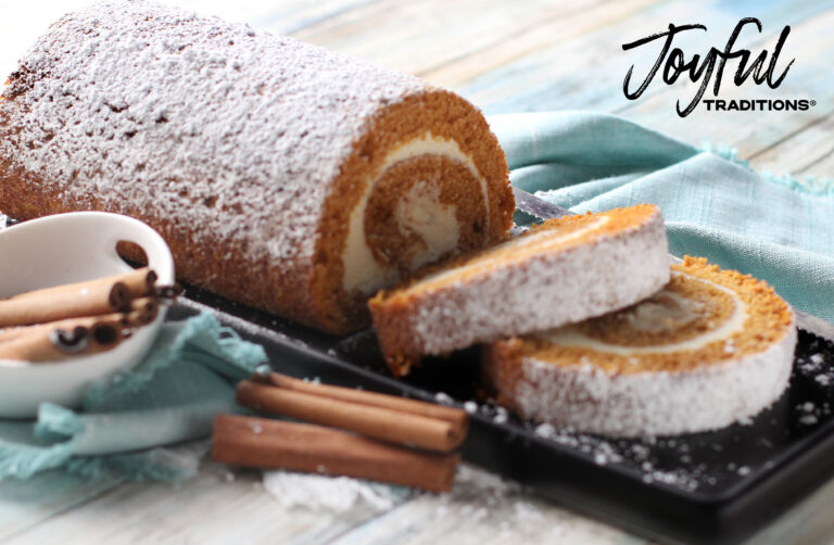 Pumpkin Cake Roll on black cutting board with blue table cloth on wooden background. Garnished with cinnamon sticks on table and in a small white bowl. Has Joyful Traditions logo.